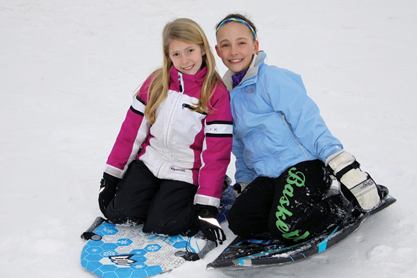 Two girls sitting on sleds on a snowy hill.