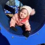 A child in a piece of playground equipment, wearing a winter coat and hat.