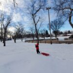 A child outdoors in a park pulling a slide on the snow.