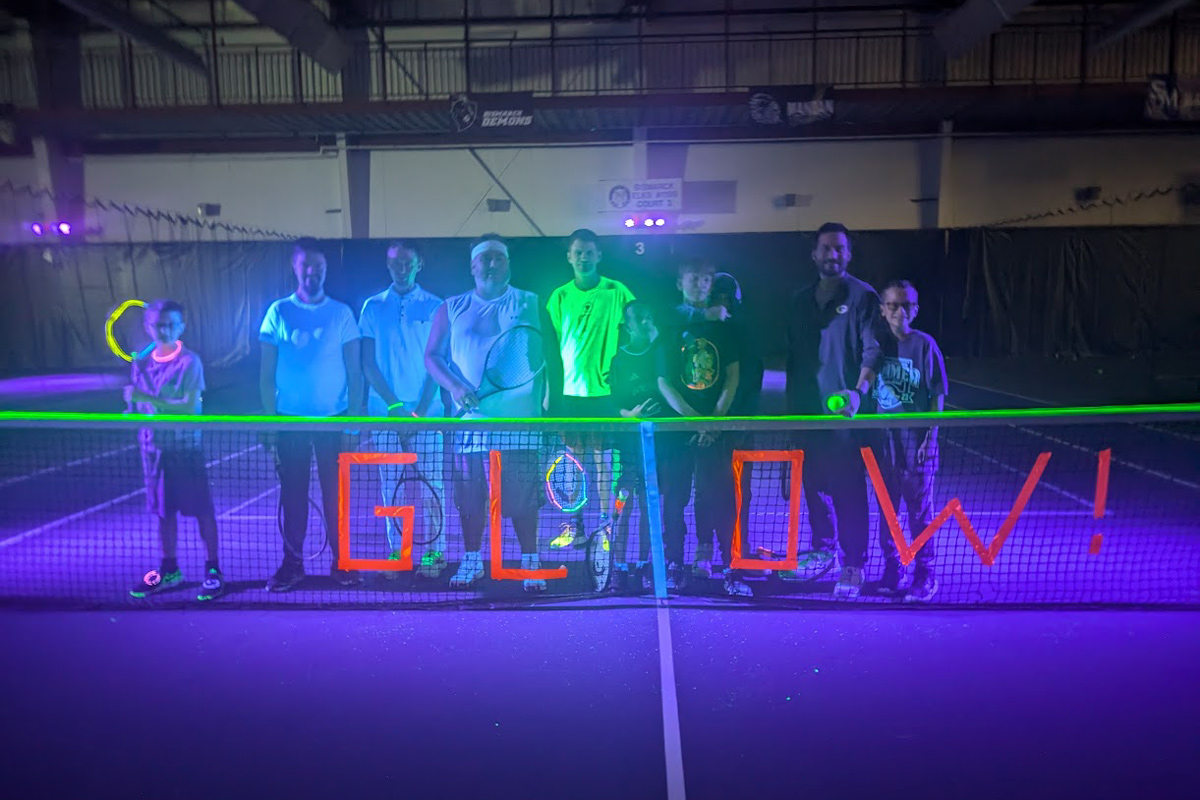 Blacklight-Tennis-2024 group photo of blacklight tennis participants in front of a tennis net that says "Glow!" in glow in the dark duct tape