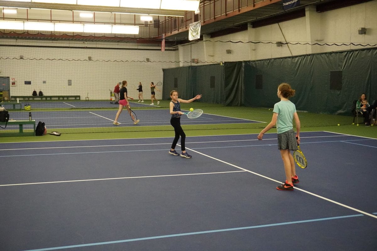 two kids playing tennis, one girl hits a tennis ball from underneath it