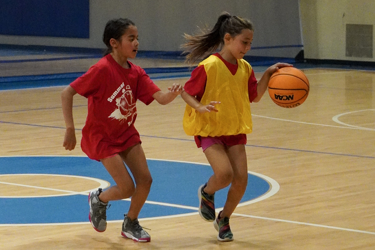Two kids playing youth basketball in a gym. One is dribbling the basketball, and the other is playing defense.