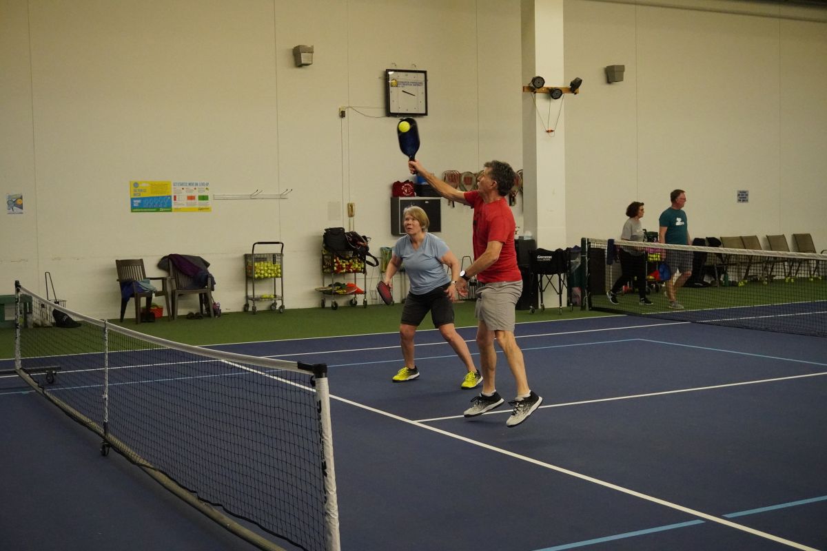 Two people playing pickleball together on an indoor court. One player puts his paddle up to hit the ball.