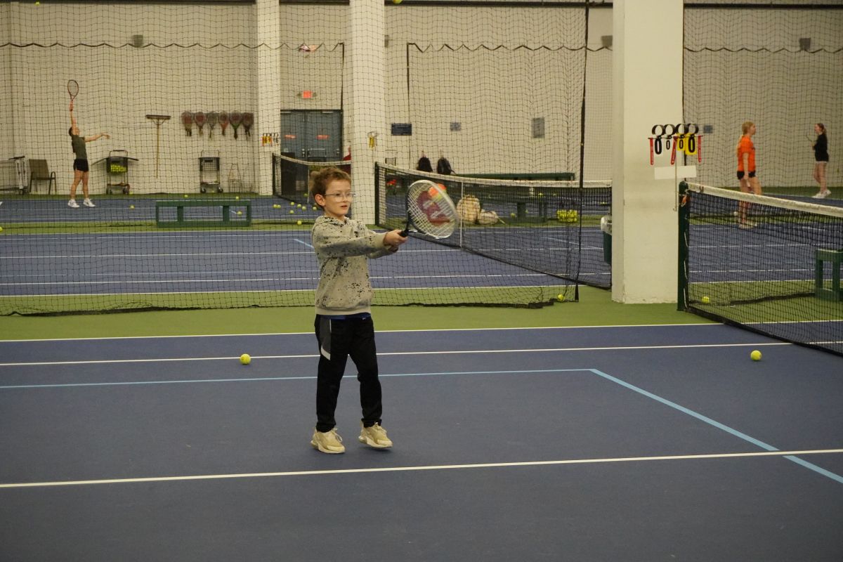 a child hits a tennis ball with his racquet at an indoor court.